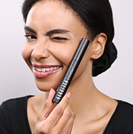 En kvinde smiler bredt, mens hun holder en mascarabørste og viser sin begejstring for skønhed og makeup-anvendelse.